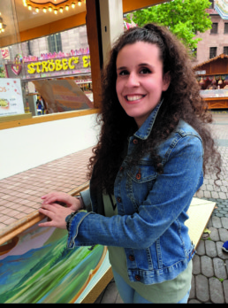 Francesca Di Nato steht vor einer Marktbude am Jakobsplatz in Nürnberg und lächelt in die Kamera.