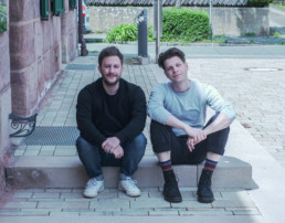 Andreas Jäger und Julian Menz sitzen auf der Treppe vor ihrem Büro