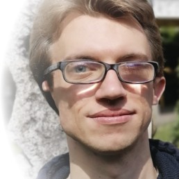 Edvin Duric mit Brille, kurzen dunkelblonden Haaren und leichten Bartstoppeln. Er trägt einen dunklen Hoodie und steht im Freien vor einem Felsen.