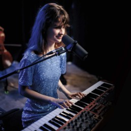 CassMae, eine junge, schlanke Frau spielt am Klavier und lächelt dabei leicht. Vor ihr steht ein Mikrofon. Sie hat lange, glatte braune Haare mit Pony und trägt ein blaues Paillettenkleid mit Puffärmeln.