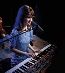 CassMae, eine junge, schlanke Frau spielt am Klavier und lächelt dabei leicht. Vor ihr steht ein Mikrofon. Sie hat lange, glatte braune Haare mit Pony und trägt ein blaues Paillettenkleid mit Puffärmeln.