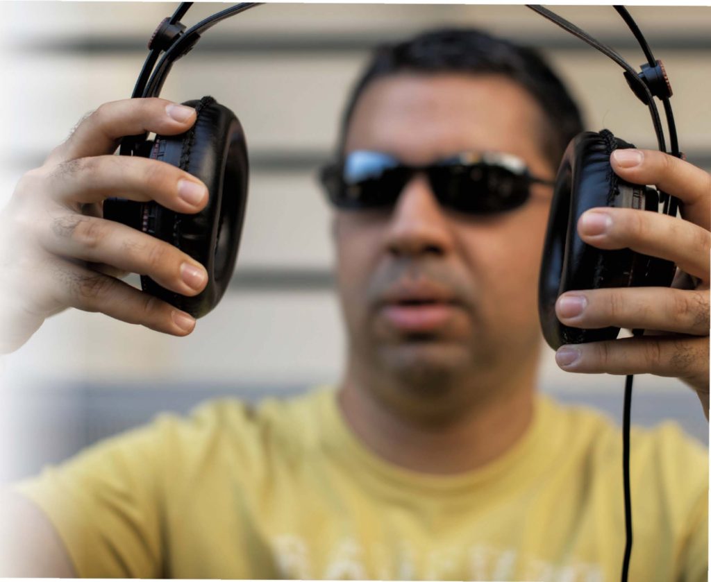 DJ Artin, ein Mann mit Dreitagebart. Er trägt eine dunkle Sonnenbrille und ein gelbes T-Shirt. Er hat große, schwarze Lederkopfhörer in beiden Händen.