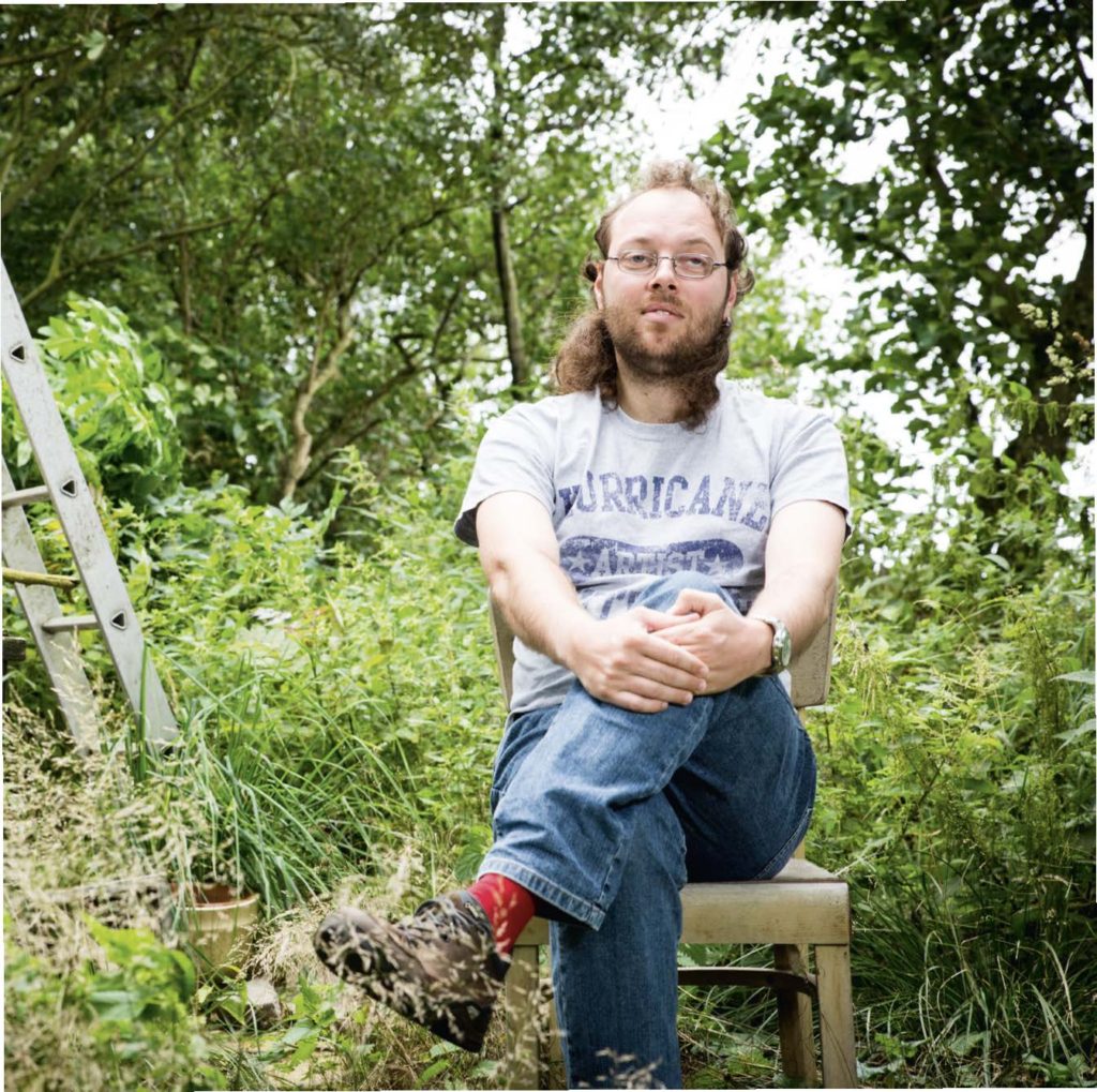 Ernesto Schnettler, der Gitarrist der Band Station 17. Er trägt ein Shirt des Hurrican-Festivals und Jeans und sitzt mit überschlagenen Beinen auf einem Hocker auf einer Wiese. Überlappend sieht man links davon ein schwarz-weiß-Foto, das seine Band auf der Bühne zeigt. 