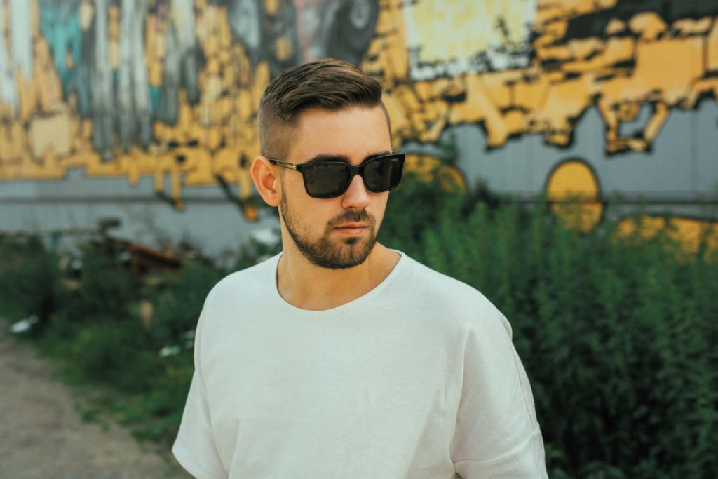 Daniel beziehungsweise der Rapper Vokuz. Er hat braune, mit Gel frisierte Haare und einen Fünftagebart. Er trägt ein weißes T-Shirt und eine schwarze Sonnenbrille. Er steht im Freien vor einer Graffiti-Wand. 