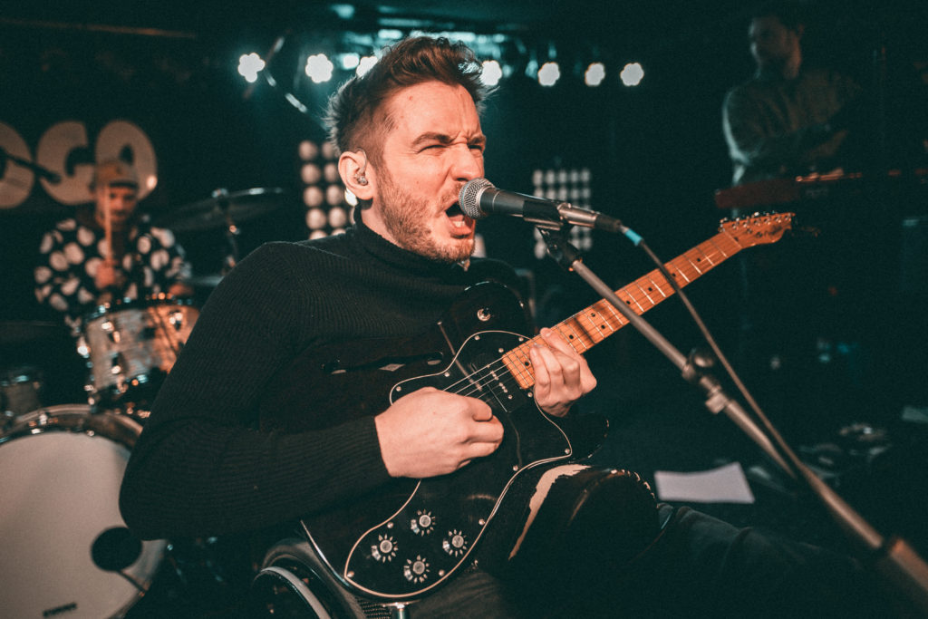 Felix Brückner mit kurzen, zur Tolle frisierten braunen Haaren in einem schwarzen Rollkragenpullover. Er sitzt im Rollstuhl auf der Bühne, seine Band ist verschwommen im Hintergrund zu sehen. Er spielt E-Gitarre und singt dynamisch in ein Standmikro.