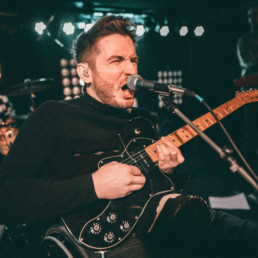 Felix Brückner mit kurzen, zur Tolle frisierten braunen Haaren in einem schwarzen Rollkragenpullover. Er sitzt im Rollstuhl auf der Bühne, seine Band ist verschwommen im Hintergrund zu sehen. Er spielt E-Gitarre und singt dynamisch in ein Standmikro.